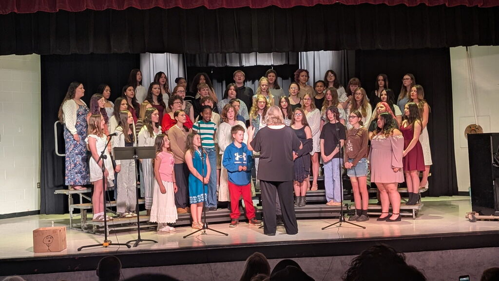 A group of students stand on risers with music stands in front of them. There is a lady in black with her back towards the audience directing them.