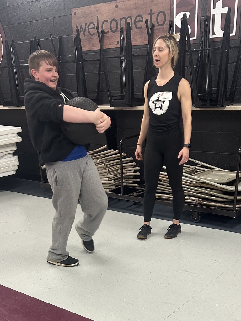 A boy is carrying a weighted ball while a lady with a warrior body tshirt talks to him. There are chairs stacked behind the lady and a counter with stools on it.