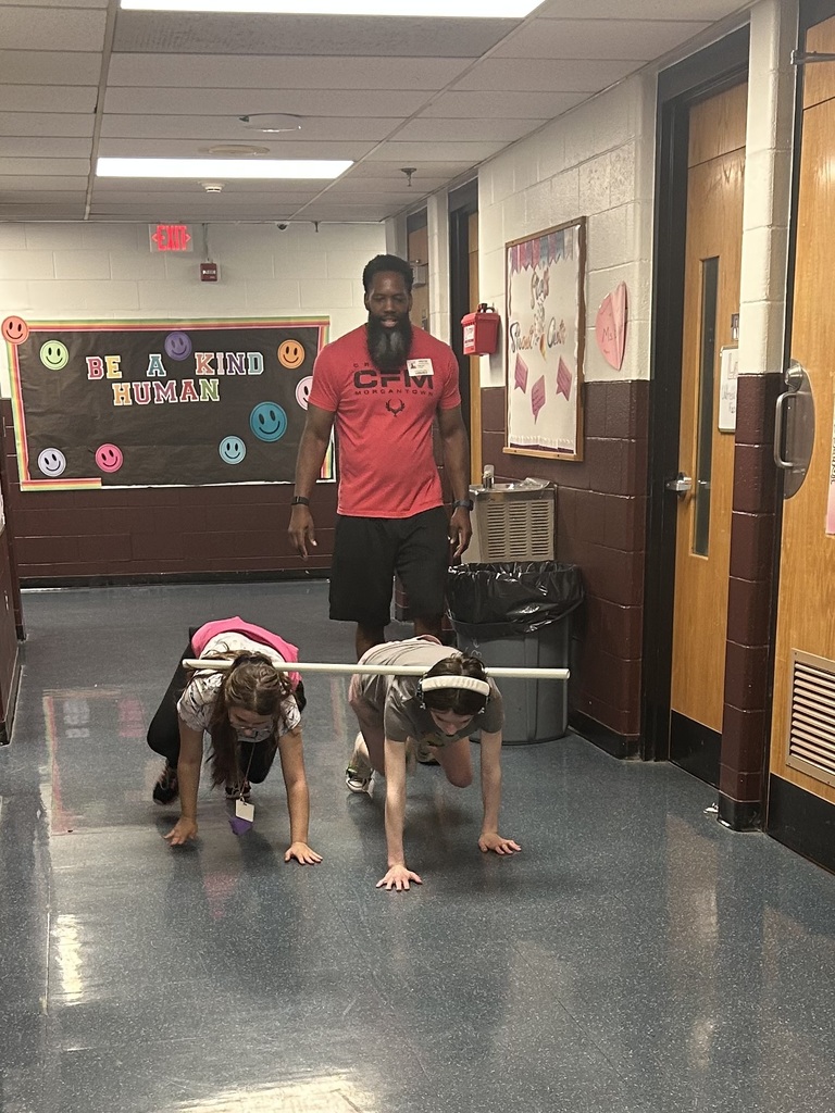 Two students are carrying a piece of white pipe across their backs as a gentleman stand behind them. They are all in a school hallway. There is a bulletin board in the background that says Be a kind human.