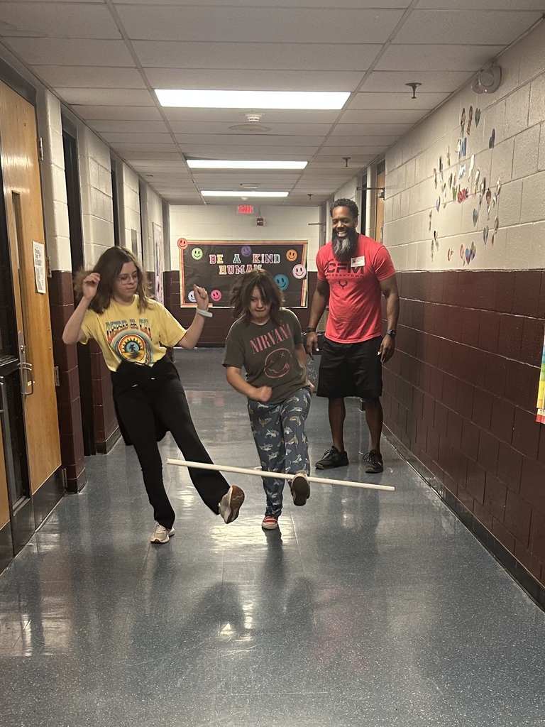Two girls are in a school hallway trying to balance a white stick on their legs. There is a gentleman behind them giving instructions.