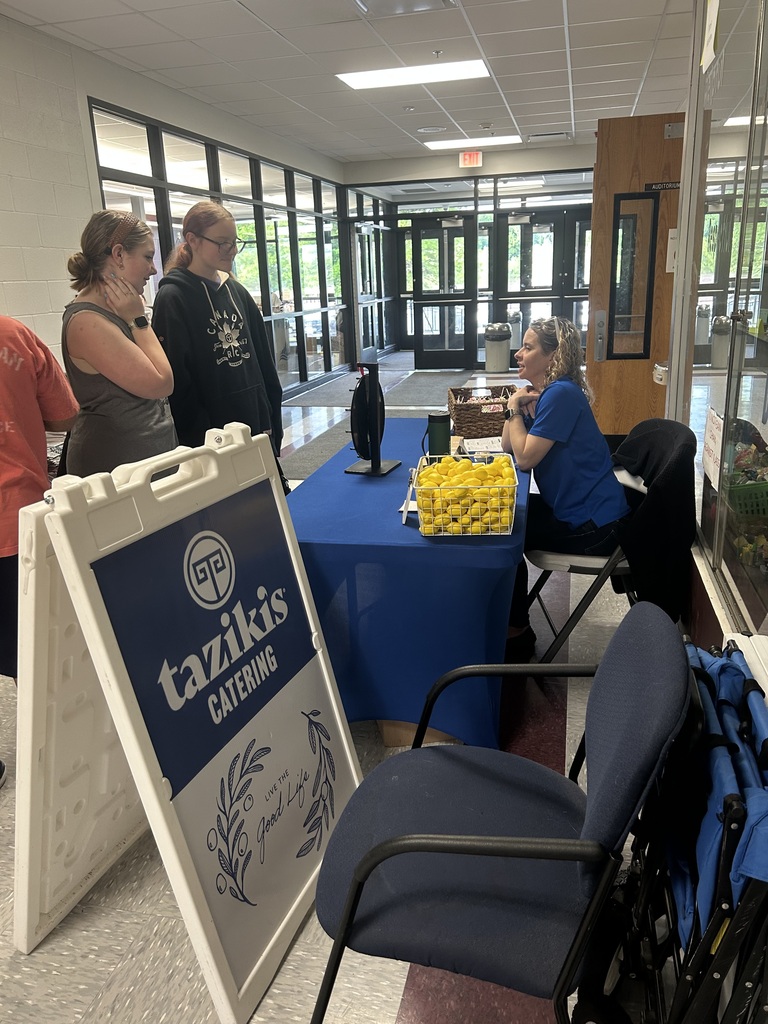 2 ladies are at a table with a spinning wheel on it and lemons. There is a lady sitting at the table beside a Tazikies Catering sign.
