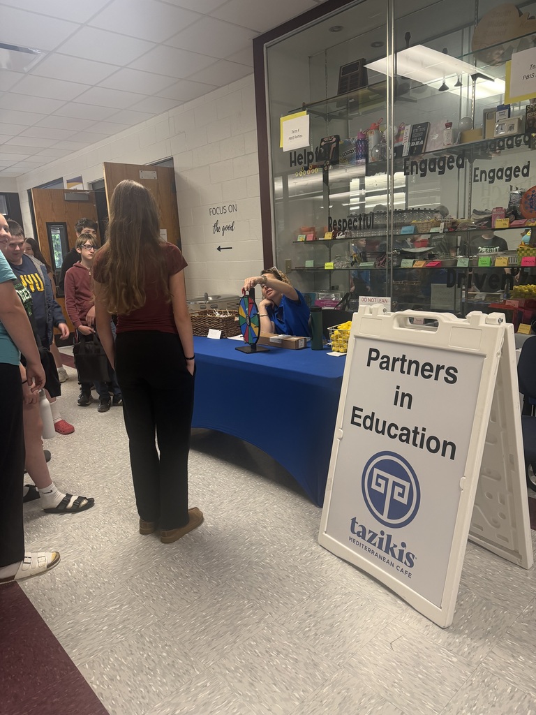 Kids standing by a blue table with a wheel on it. There is a sign saying Partners in Education Tazikis on it. The table is sitting in a hallway with a glass cabinet behind it.
