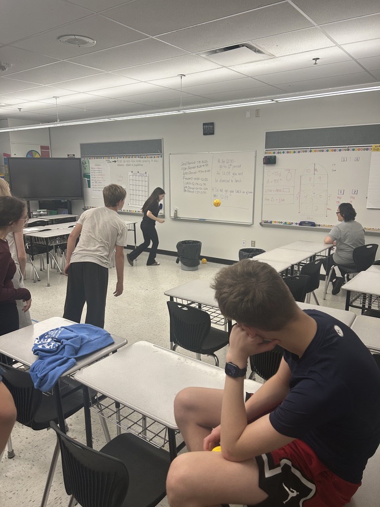Students in a classroom throwing balls at 2 garbage cans.