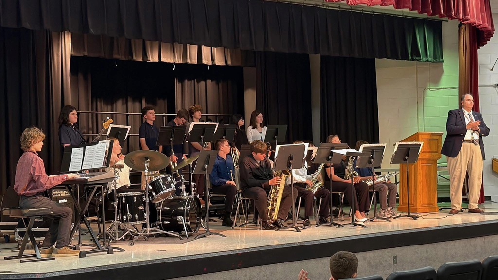 Band students are sitting on the stage in an auditorium. There is a podium with a gentleman standing beside it. There are students really to play music.