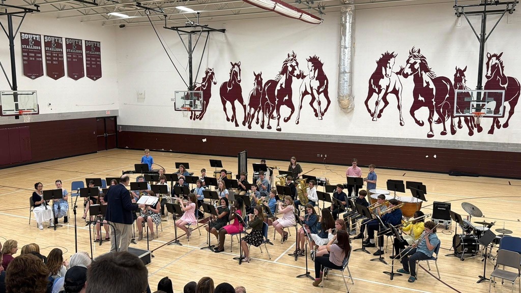 A group of band students are sitting in a gym with music stands in front of them. There are stallions painted on the back gym wall.