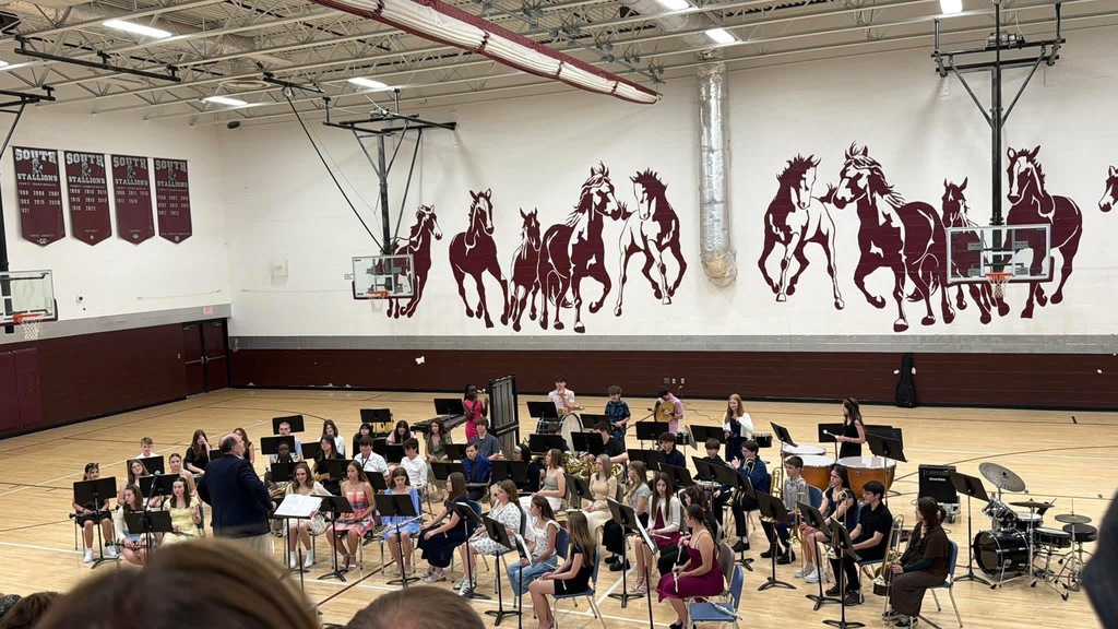A group of band students are sitting in a gym with stallions painted on the back wall. There are music stands on the floor in front of the students.