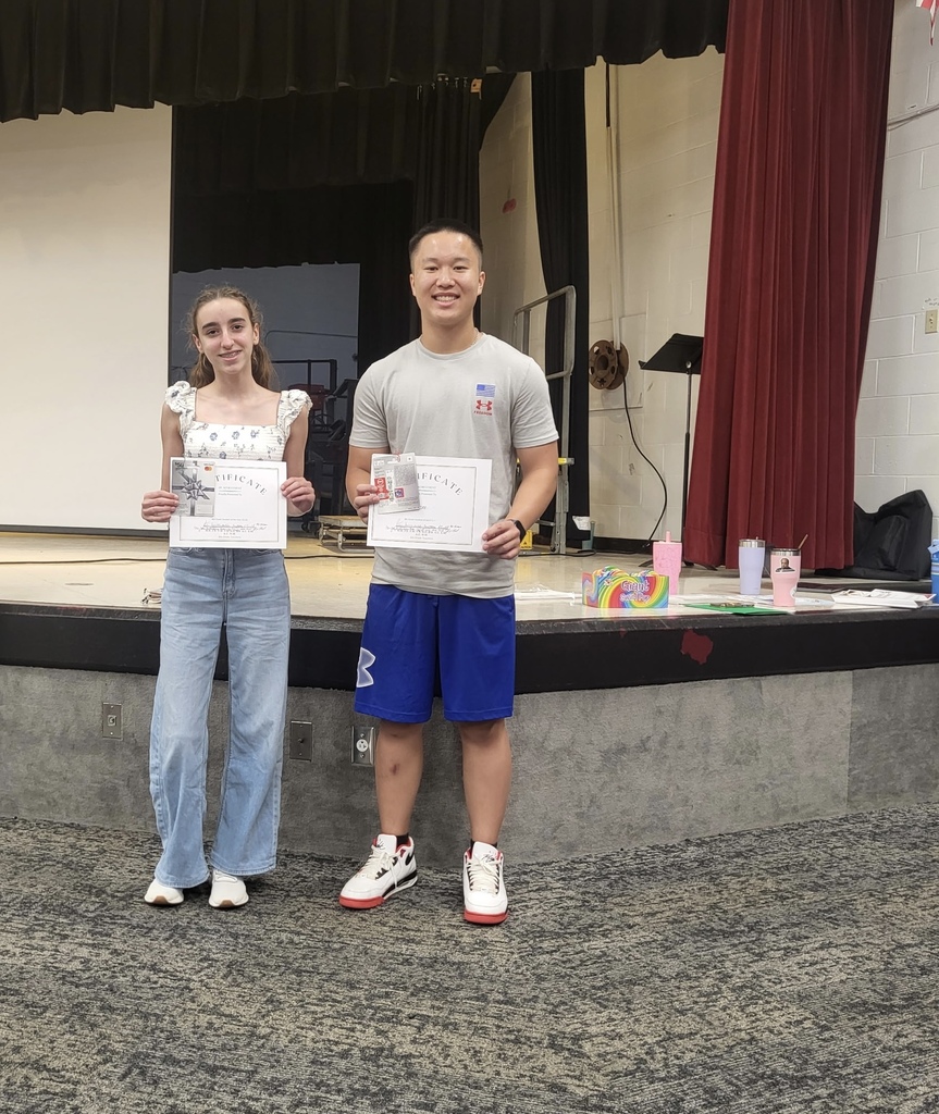 2 students standing near a stage holding up certificates and gift cards. One student is wearing jeans and a short sleeve shirt and the other student is wearing blue shorts and a grayish t-shirt. Both students are smiling.