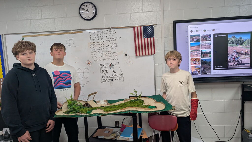 Three boys standing beside a project in a classroom. There is a TV screen with photos on it in the background.