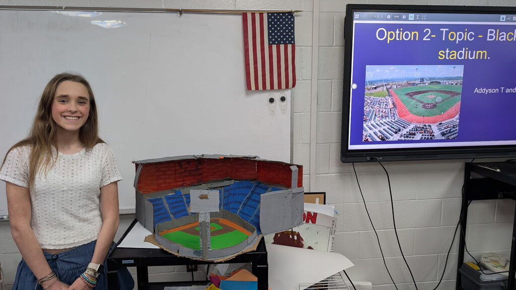 A girl is standing beside a field project in a classroom. There is a TV screen in the background with a stadium presentation on it.