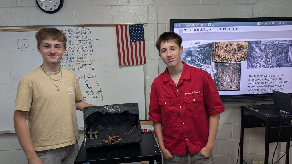 Two boys standing beside a project in a classroom. There is a TV screen with photos and words in the background.