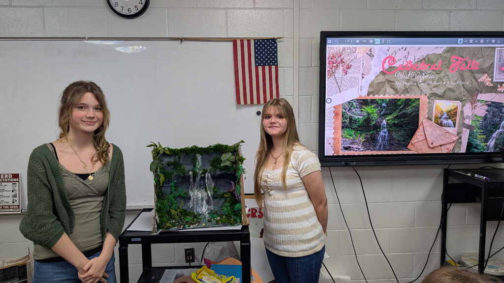 Two girls standing beside a project of a waterfall in a classroom. There is a TV screen in the background with a slideshow on it.