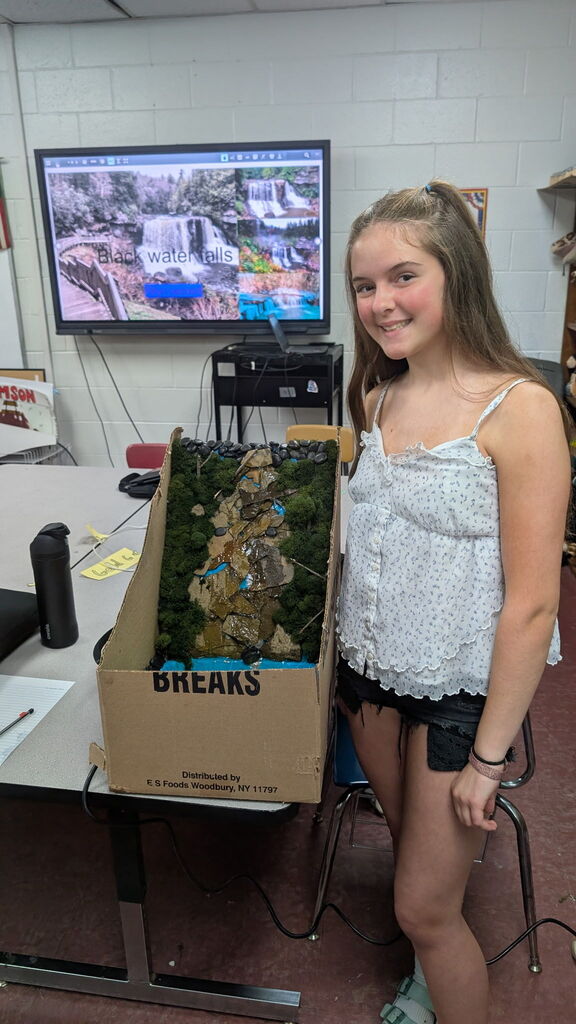 A girl standing in a classroom beside a Blackwater Falls Project in a cardboard box. There is a screen with a picture of Blackwater Falls.