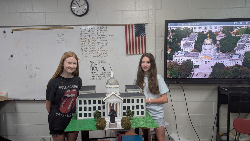 Two girls standing beside a project of a white building. The building is also on a TV screen. This is in a classroom.
