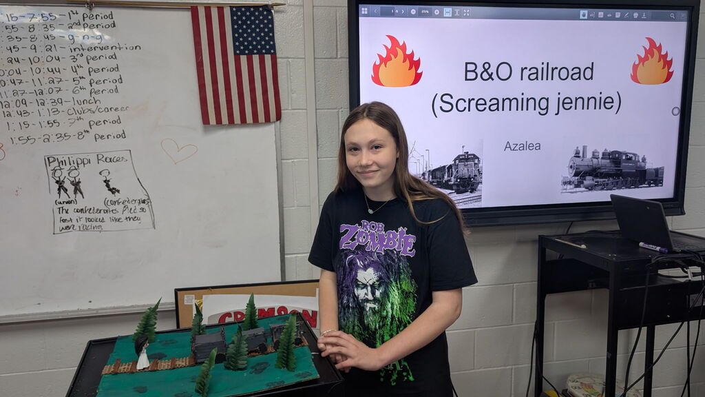 A girl standing beside a project in a classroom. There is a screen in the background with B&O Railroad and 2 photos of trains.