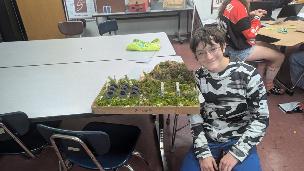 A boy sitting in a classroom beside a box with a project in it.