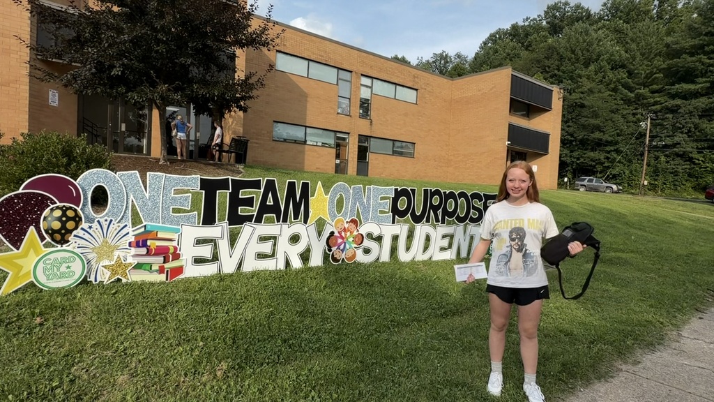 A girl holding a Chromebook case and a piece of paper standing in front of South Middle School. There is a sign that says One Team - One Purpose - Every Student. There is a car beside the building in the background.
