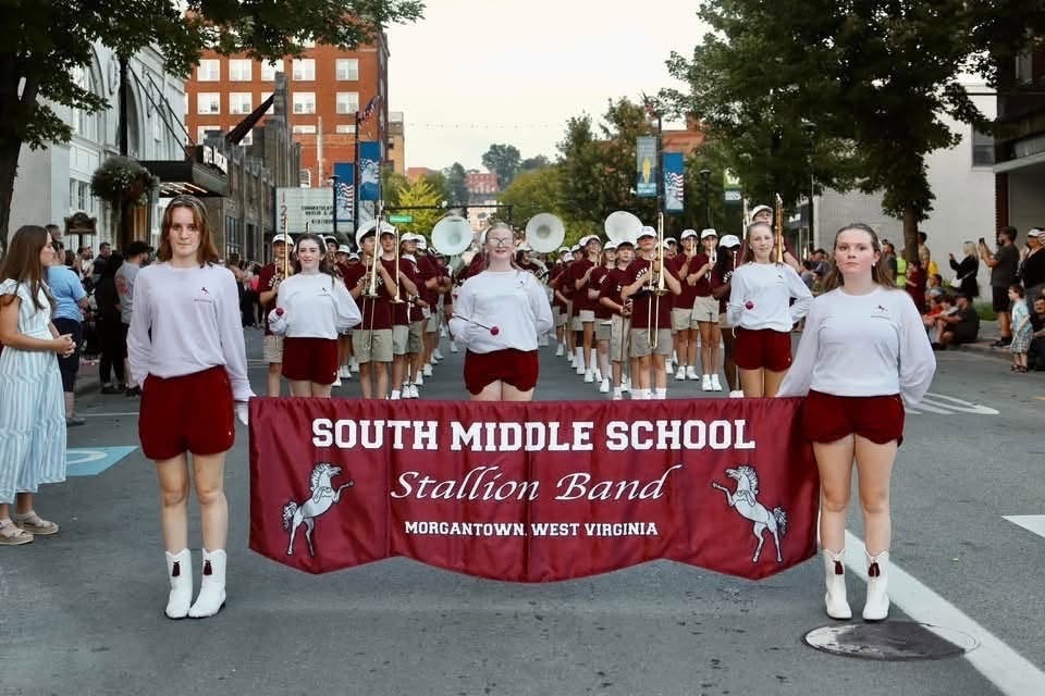 2 girls holding a South Middle School Stallion Band banner in front of a marching band. The band is standing in the middle of the road in a parade formation.