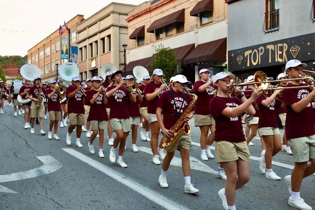 A bunch of band members dressed in tan shorts, maroon Stallion Band shirts, white tennis shoes, and white hats. They are holding their instruments up, marching, and playing their instruments in what appears to be a parade.