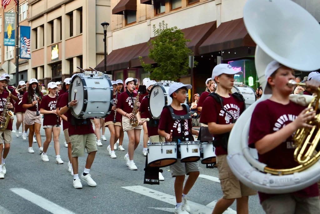 A bunch of band members dressed in tan shorts, maroon Stallion Band shirts, white tennis shoes, and white hats. They are holding their instruments up, marching, and playing their instruments in what appears to be a parade.