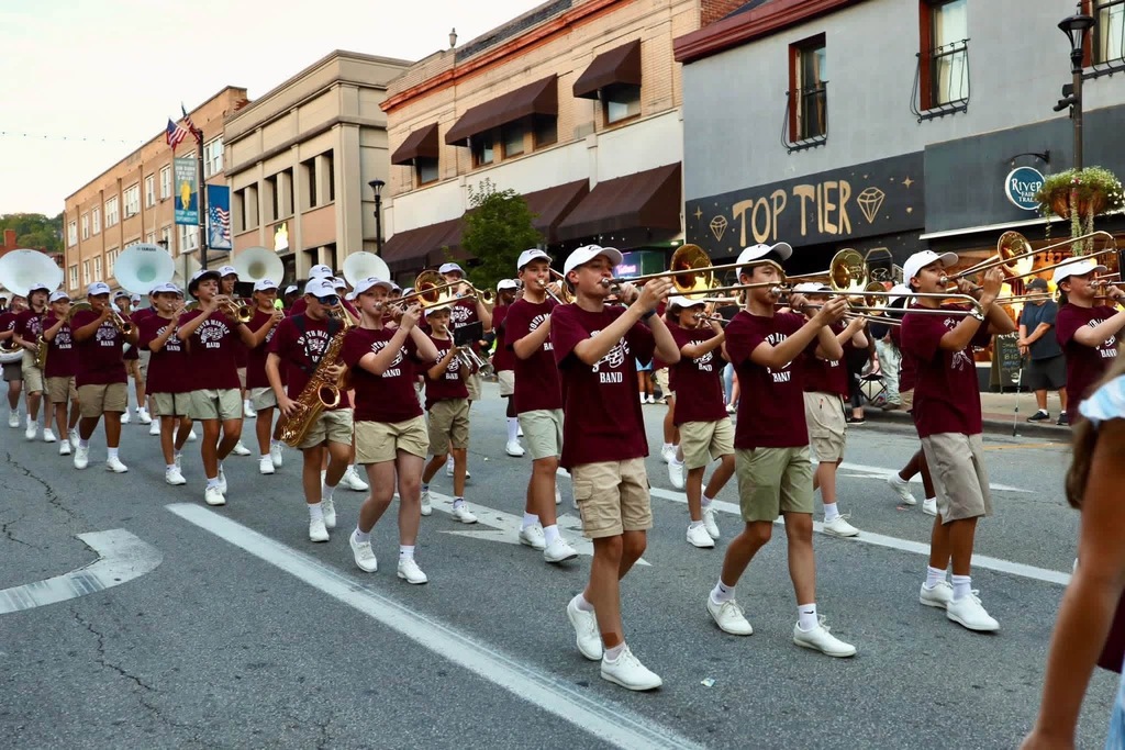 A bunch of band members dressed in tan shorts, maroon Stallion Band shirts, white tennis shoes, and white hats. They are holding their instruments up, marching, and playing their instruments in what appears to be a parade.