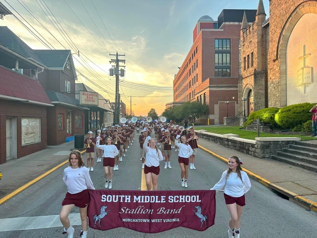 2 girls holding a South Middle School Stallion Band banner in front of a marching band. The band is standing in the middle of the road in a parade formation. There is a church on the right and a business on the left.