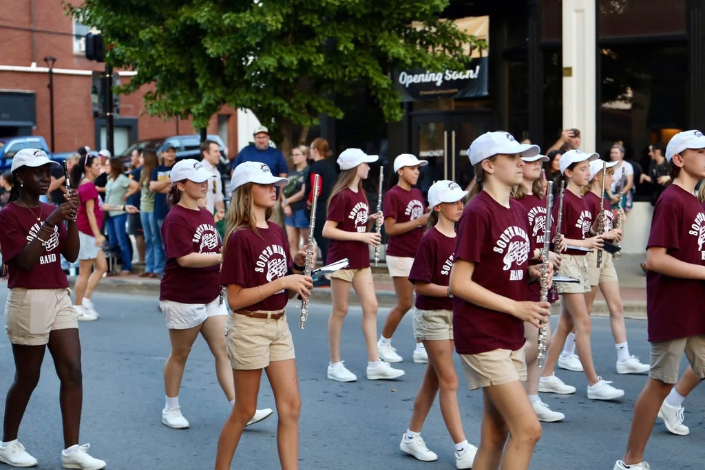 A bunch of band members dressed in tan shorts, maroon Stallion Band shirts, white tennis shoes, and white hats. They are holding their instruments up and marching  in what appears to be a parade.