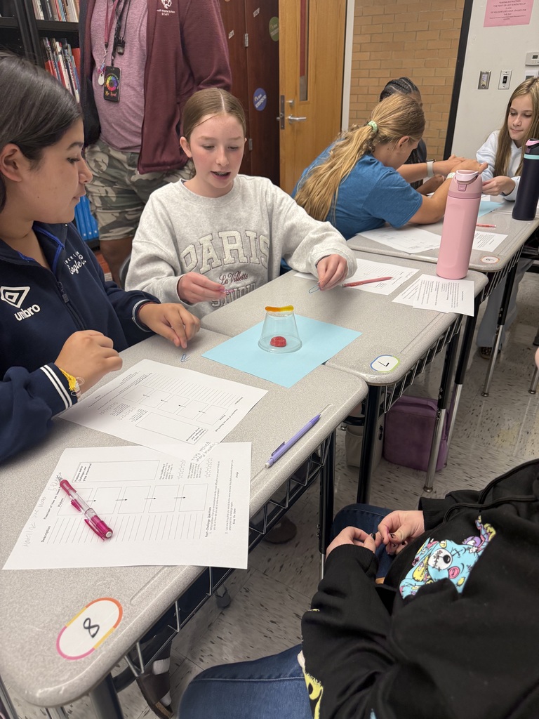 Students sitting in a classroom at desks. There is a cup turned over with a red thing inside the cup. Students have papers sitting on desks.