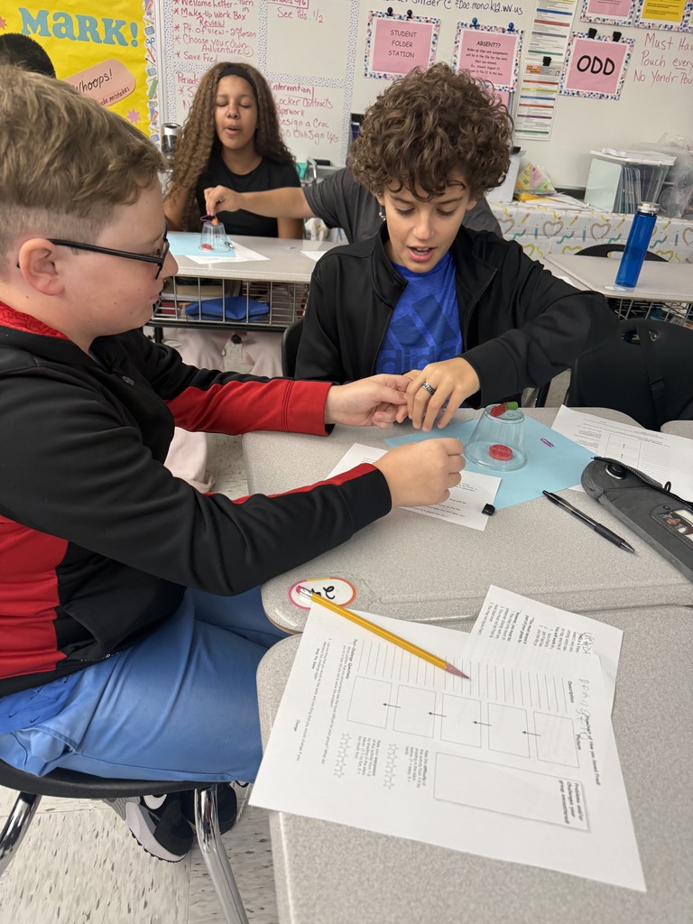 Students sitting in a classroom at desks. There is a cup turned over with a red thing inside the cup. Students have papers sitting on desks.