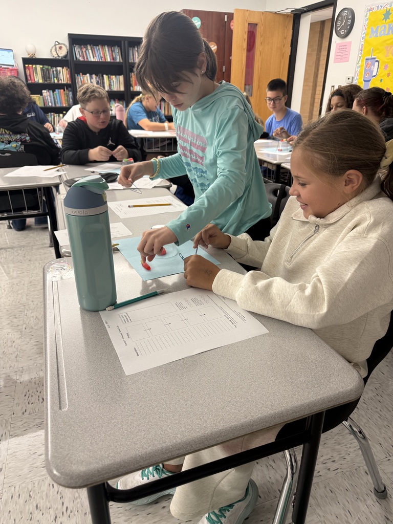Students are sitting at desks in a classroom. Two students are working together and have papers on their desks. Other students at desks are in the background.