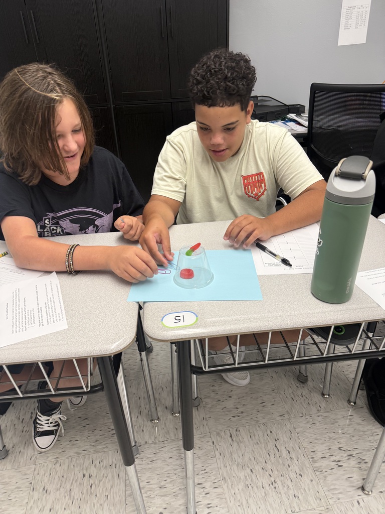 Students sitting in a classroom at desks. There is a cup turned over with a red thing inside the cup. Students have papers sitting on desks.