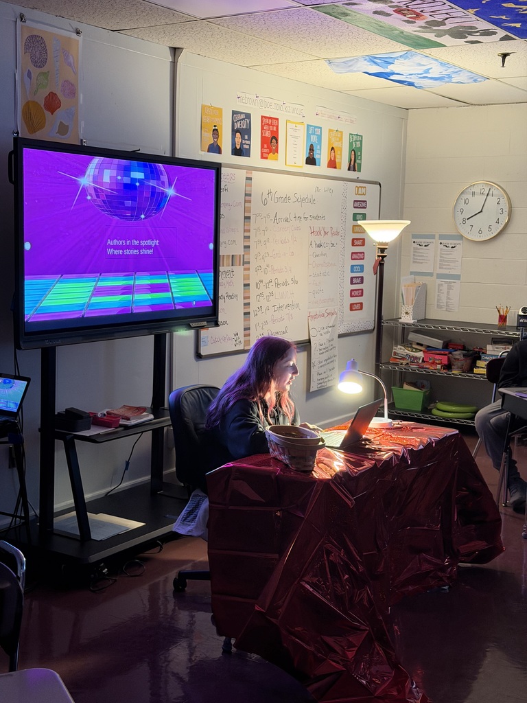 Student sitting at a desk in a dark room with 2 lights shining on them. There is a white board behind and students are sitting at desks.