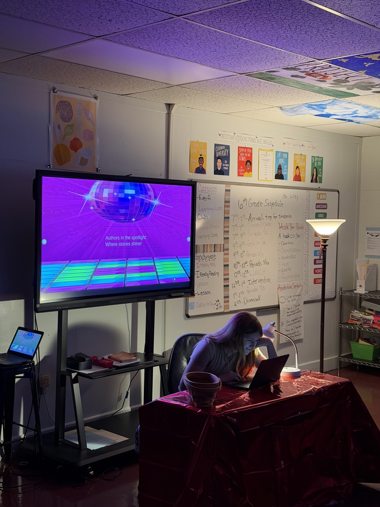 Student sitting at a desk in a dark room with 2 lights shining on them. There is a white board behind and students are sitting at desks.