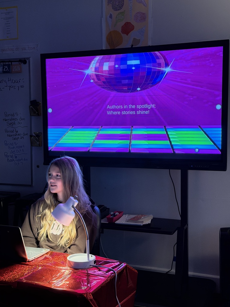 Student sitting at a desk in a dark room with 2 lights shining on them. There is a white board behind and students are sitting at desks.