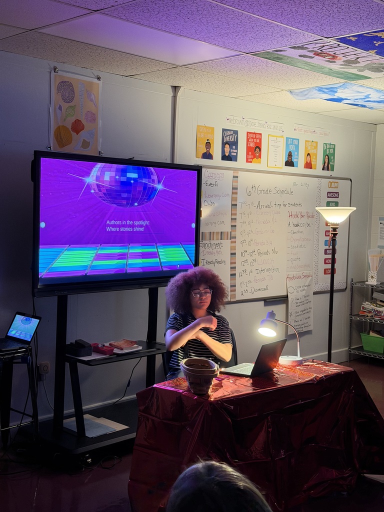 Student sitting at a desk in a dark room with 2 lights shining on them. There is a white board behind and students are sitting at desks.