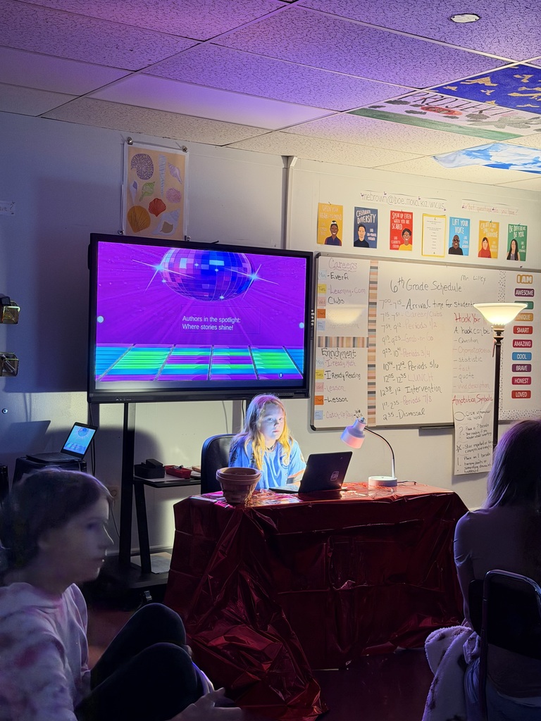 Student sitting at a desk in a dark room with 2 lights shining on them. There is a white board behind and students are sitting at desks.