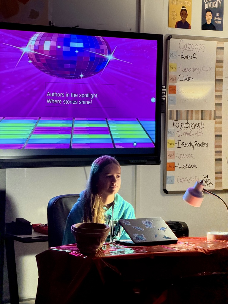 Student sitting at a desk in a dark room with 2 lights shining on them. There is a white board behind and students are sitting at desks.