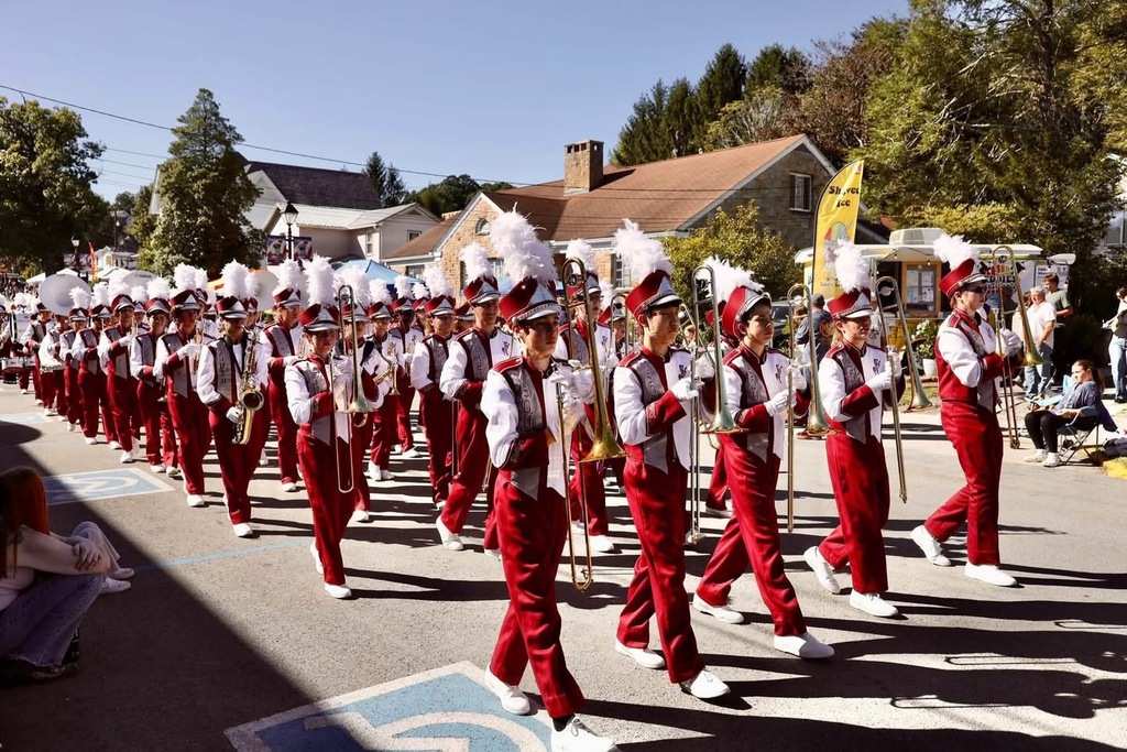 A band marching and playing instruments in a parade. The band members are wearing white and marroon uniforms and have a white feather coming out of their hats. People are along the road watching the parade.