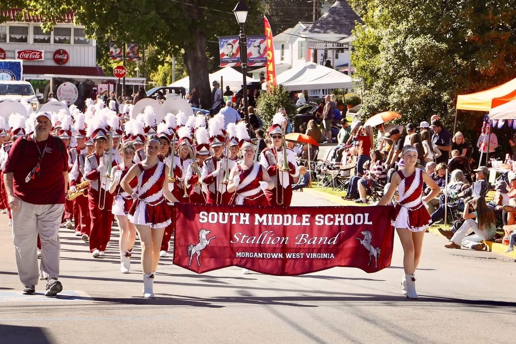 A band wearing maroon and white uniforms. A man walking beside 2 girls carrying a banner that says South Middle School Stallion Band Morgantown, WV. They are walking down a street in a parade while spectators are along the road watching.