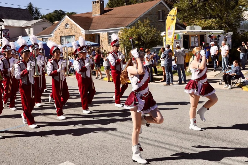 A band marching and playing instruments in a parade. The band members are wearing white and marroon uniforms and have a white feather coming out of their hats. People are along the road watching the parade.