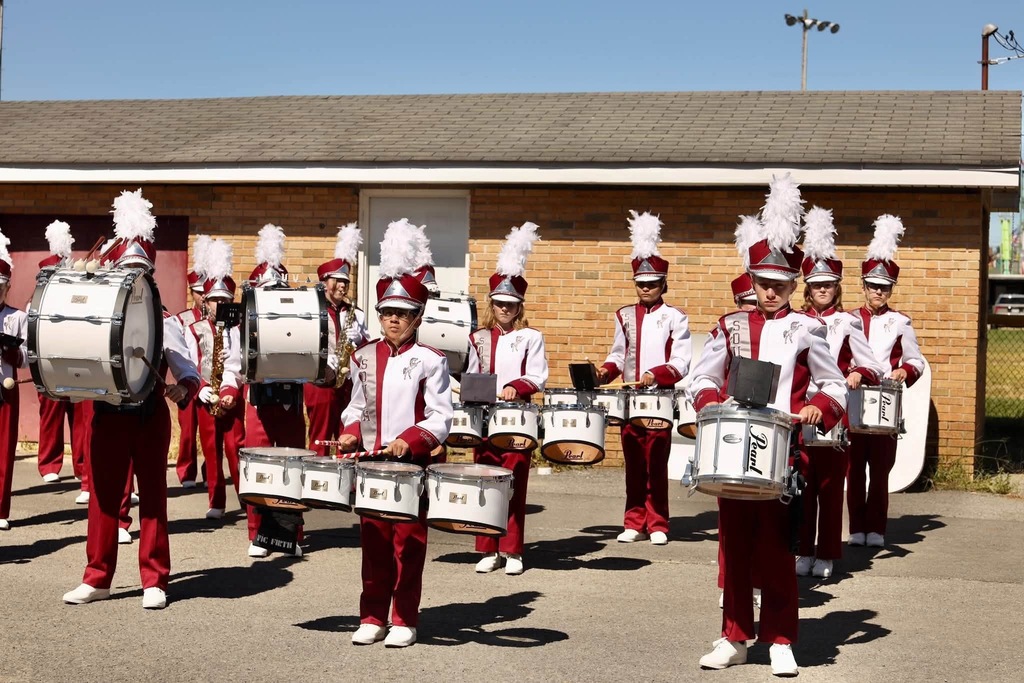 Band members holding drums outside in a parking lot. There is a building in the background and the drummers are wearing marroon and white band uniforms.