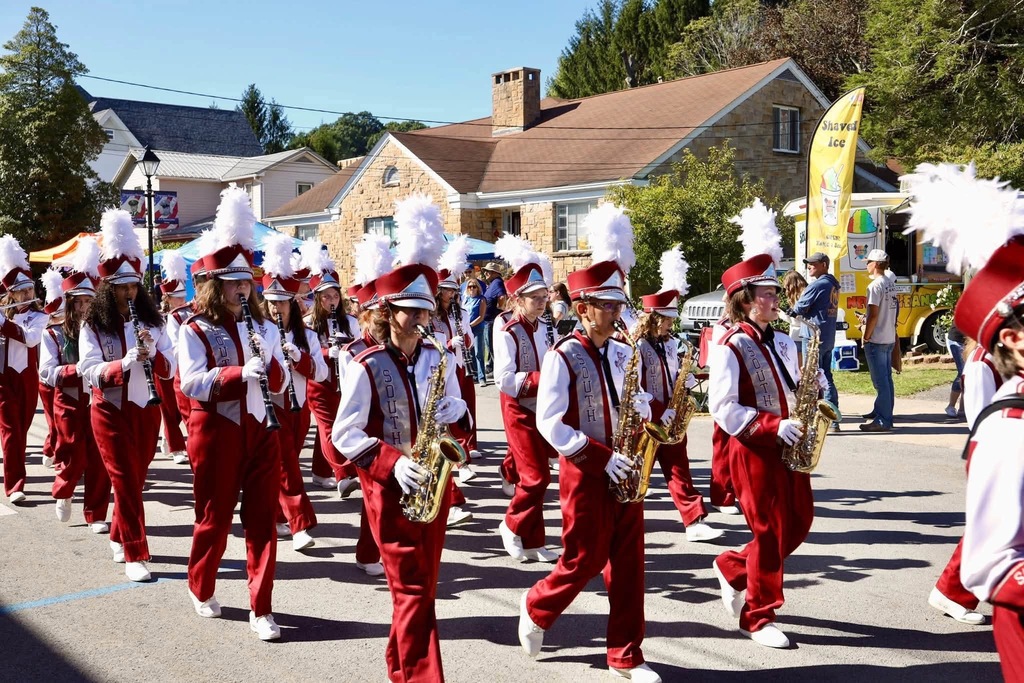 A band marching and playing instruments in a parade. The band members are wearing white and marroon uniforms and have a white feather coming out of their hats. People are along the road watching the parade.