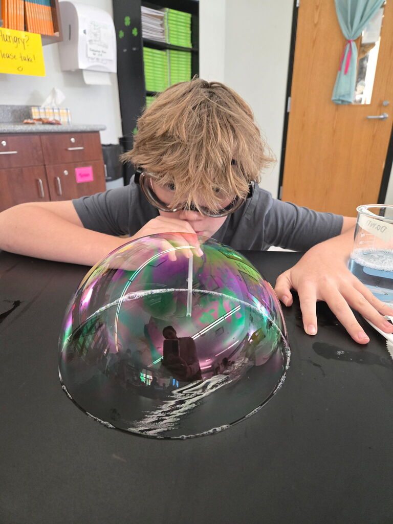 Student sitting a table blowing a bubble with a straw. 