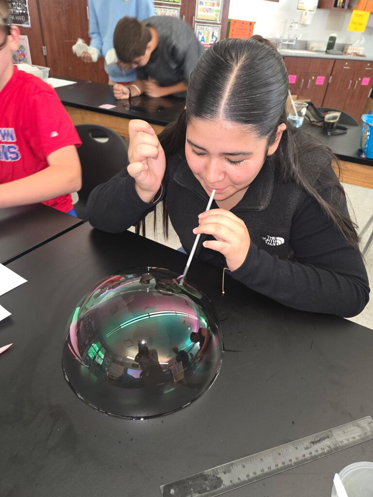 Student sitting a table blowing a bubble with a straw. A ruler is beside her.
