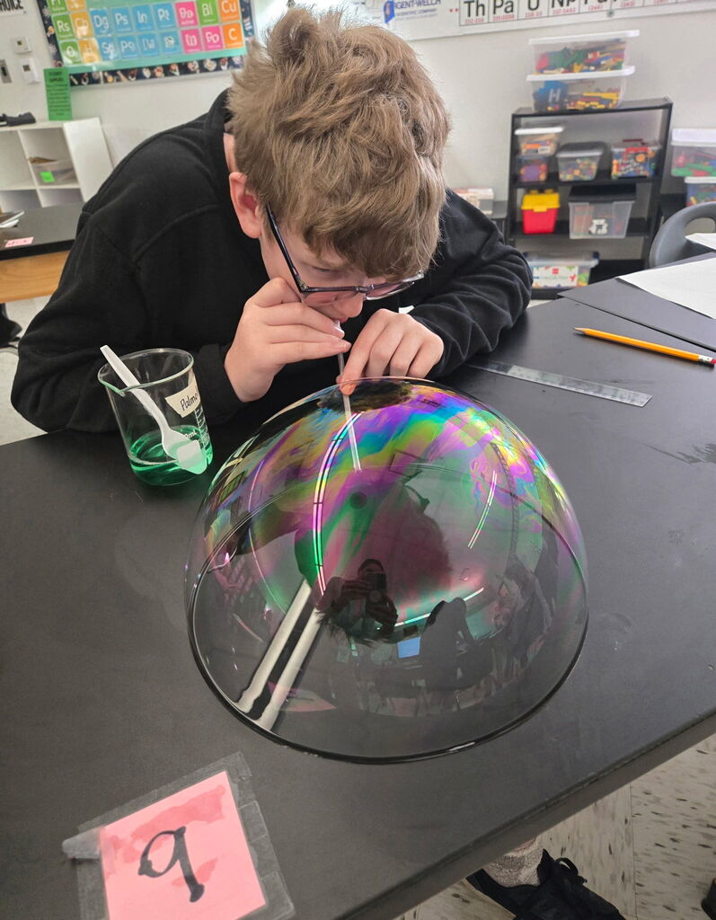 Student sitting a table blowing a bubble with a straw. A cup of soap is beside him and a number 9 is on the table.