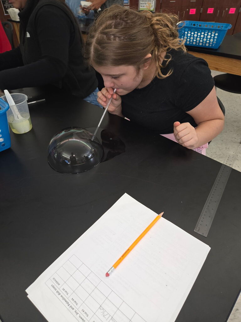 Student sitting a table blowing a bubble with a straw. A cup of soap is beside her and a sheet of paper and pencil are  on the table.