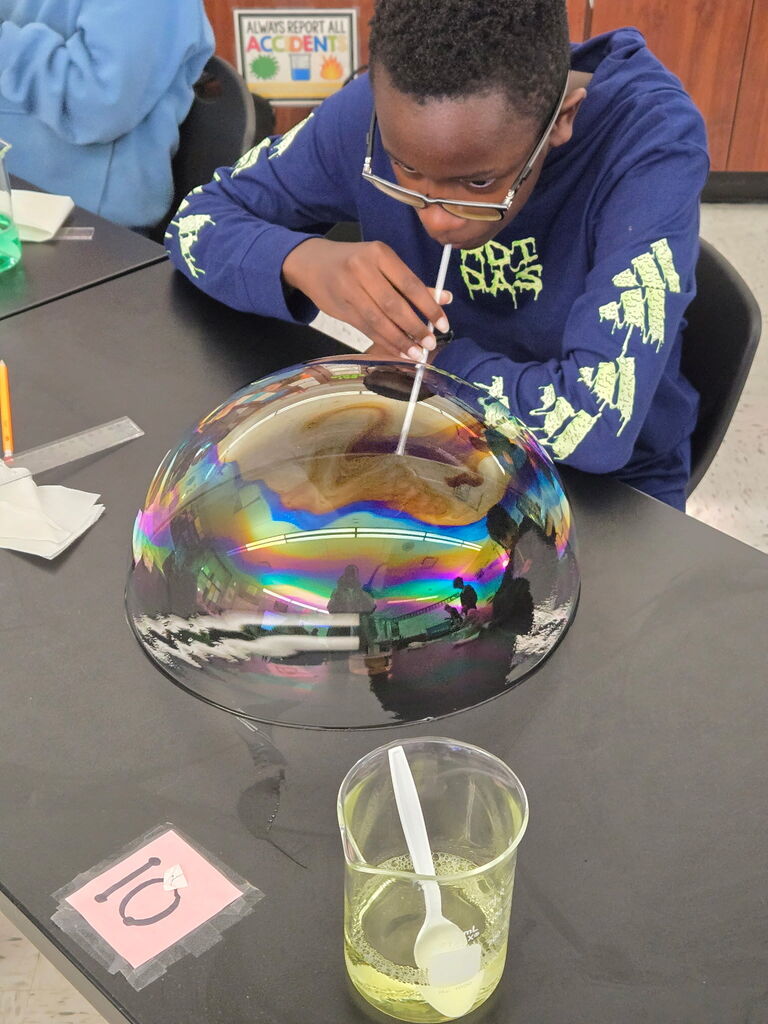 Student sitting a table blowing a bubble with a straw. A cup of soap is beside him and a number 10 is on the table.