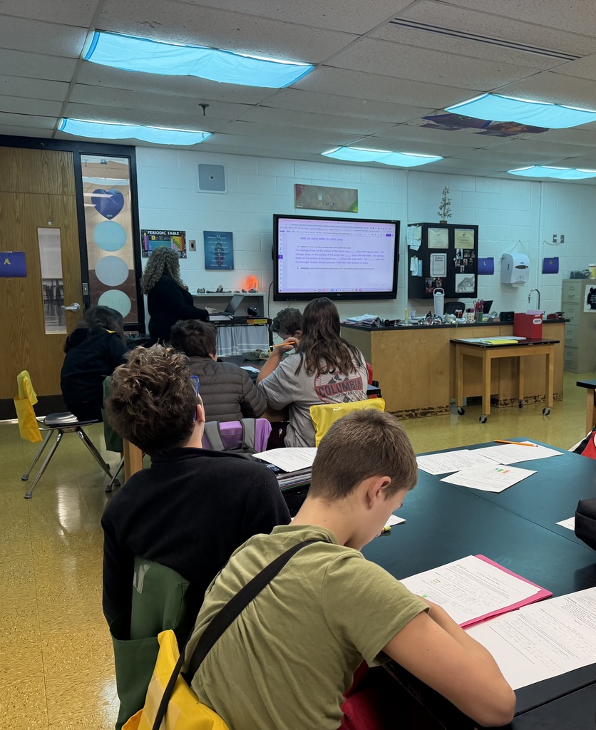 Students are in a classroom facing a teacher who is standing near a whiteboard. The students are sitting at tables and there are papers on the tables.