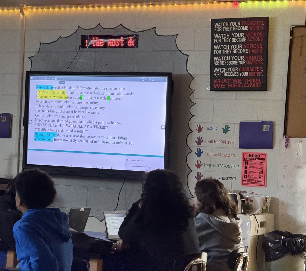 Three students in a science classroom facing a whiteboard with Science information on it. All the students have their backs to the camera and you can see 2 opened Chromebooks.