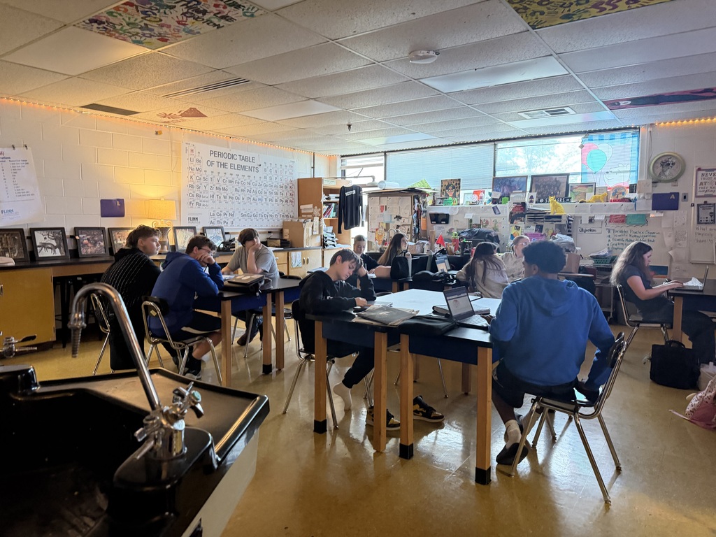 Students sitting at tables in a science classroom. Some are looking towards the right. One student has his Chromebook on a table and is looking at it. 
