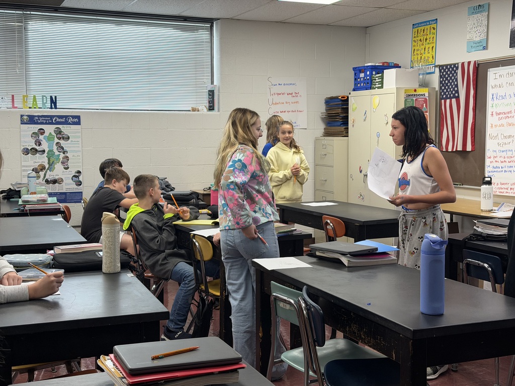 Students in a classroom. 3 are sitting at a table and 3 girls are standing. One girl has a paper in her hand. You can see 2 waterbottles sitting on black tables in a classroom.